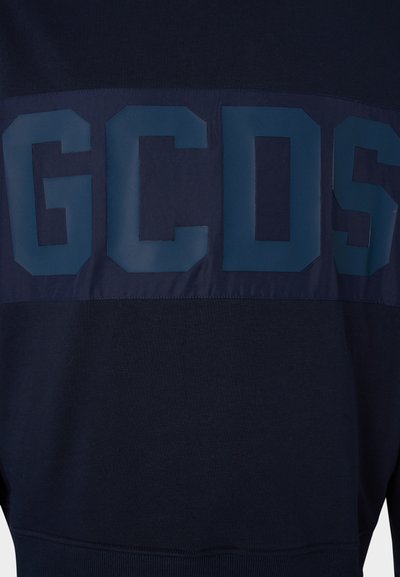 Jūras spēku džemperis ar spīguļojošu zilu paneļa daļu un lieliem burtiem "GCDS" krūtīs. Mīksta tekstūra, brīva piegriezuma un ribota apģērba maliņas.