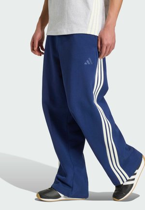 Osoba nosząca granatowe spodnie dresowe Adidas z białymi paskami po bokach oraz czarno-białe trampki, idąca.