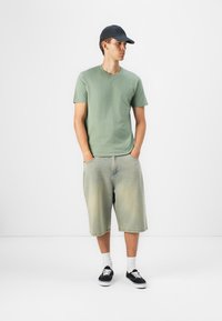 Tricou verde din bumbac, pantaloni scurți din denim bleu deschis, adidași negri și o șapcă închisă. Modelul stă cu mâinile în buzunare, pe un fundal simplu.