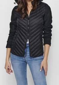 Femme portant une veste matelassée noire avec une fermeture éclair devant et un motif tressé, assortie à un jean bleu clair.