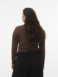 Frau mit langen, welligen braunen Haaren, die ein braunes langärmliges Oberteil und schwarze Hosen trägt, stehend mit dem Rücken zur Kamera vor einem schlichten weißen Hintergrund.