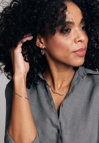 Srebrzysta, falista kolia i bransoletka noszone przez osobę o kręconych włosach, ubraną w szaro koszulę z guzikami i podwiniętymi rękawami.