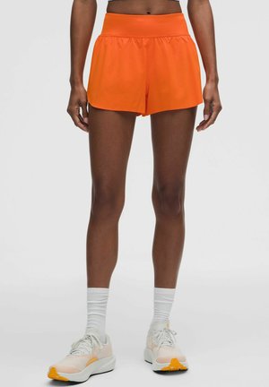 Persona que lleva puestos pantalones cortos deportivos de color naranja brillante, calcetines blancos y zapatillas de running blancas y naranjas, de pie frente a un fondo claro y liso.