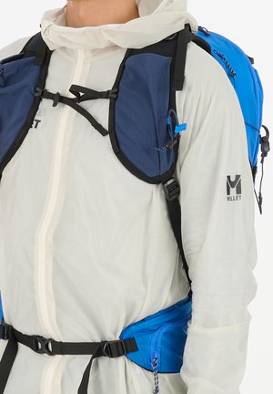 Person trägt weiße Millet-Jacke mit blauen und schwarzen Rucksackriemen und Hüftgurt, sichtbar vor weißem Hintergrund.