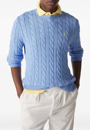 Mann trägt einen hellblauen kabelgestrickten Pullover über einem gelben Hemd mit Kragen, weiße Hose, Hände in den Taschen, vor weißem Hintergrund.