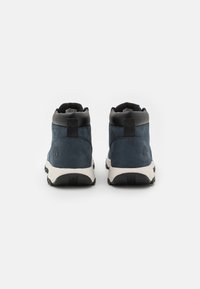 Buty mid-top z granatowego zamszu, czarną skórzaną cholewką w okolicy kostki oraz białą podeszwą. Posiadają wyraźne logo na zewnętrznej stronie pięty.