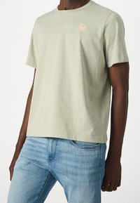 Ljusgrön t-shirt med korta ärmar, tillverkad av mjukt material, med en liten beige blomsterdesign på vänster bröst. Matchas med blå jeans.
