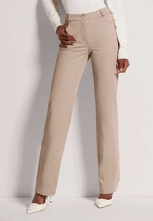 Vrouw draagt beige high-waist rechte broek met voorzakken en witte puntige hakken, staand tegen een egale muur.