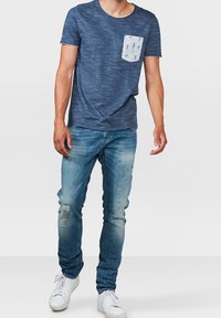 Mörkblå kortärmad t-shirt med ett mönstrat ficka, i kombination med ljusblå slitna jeans och vita sneakers.