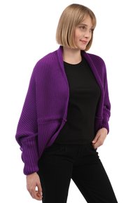 Cardigan de punto grande en color púrpura con textura acanalada, de apertura frontal y mangas largas, llevado sobre una camiseta negra y combinado con pantalones negros.