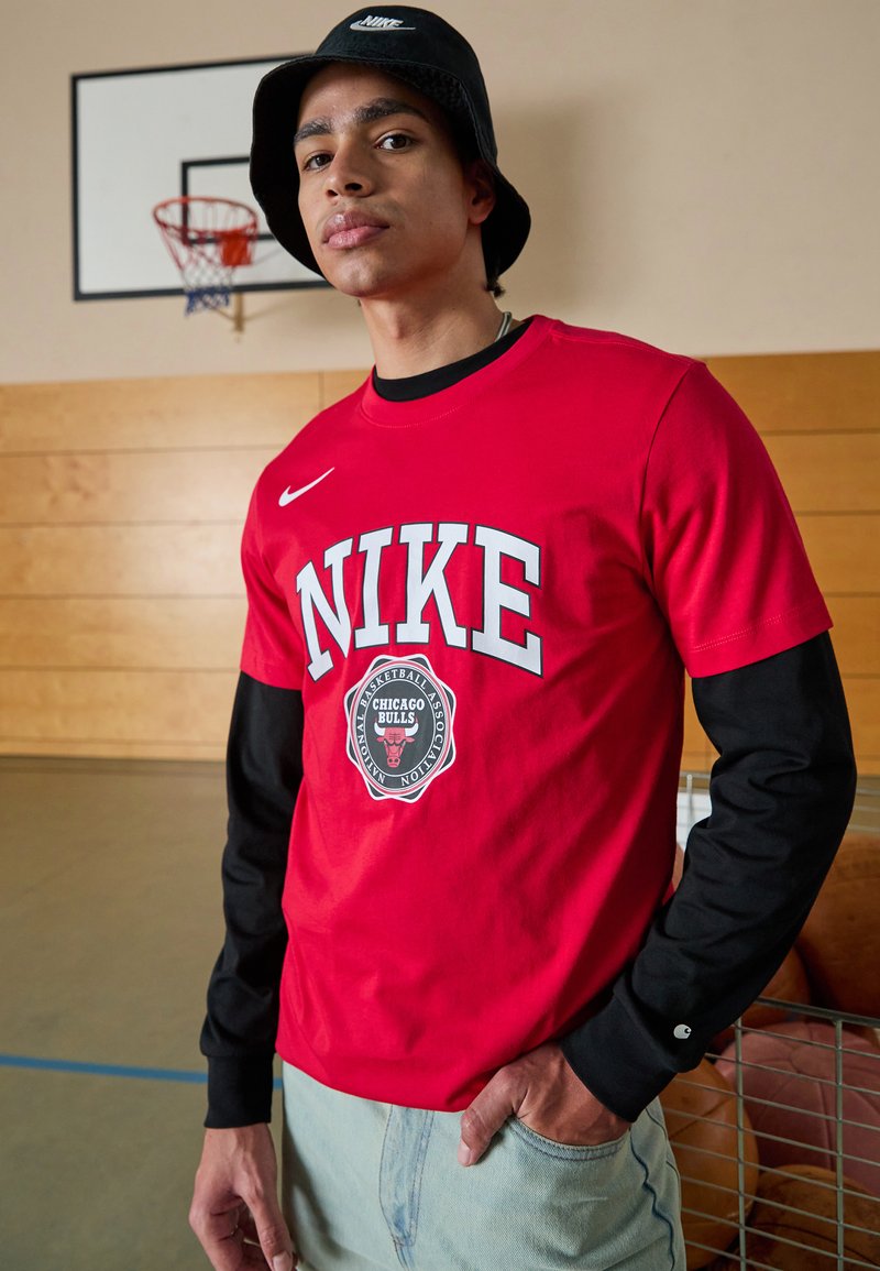 Punane lühikeste varrukatega Nike särk, millel on suur valge "NIKE" tekst, Chicago Bullsi logo ja mustad pikad varrukad. Kandmine helesiniste teksadega.