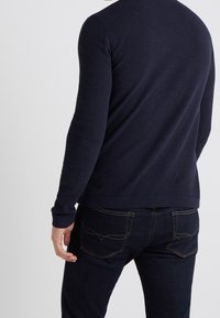 Laivastonsininen pitkähiha paita, joka on valmistettu teksturoidusta kankaasta ja jossa on pyöreä pääntie sekä istuvat hihat. Yhdistetty vaaleilla saumoilla varustettuihin tummiin farkkuihin.