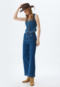 Gilet di jeans con bottoni, abbinato a pantaloni in denim a gamba larga, entrambi di un blu intenso. Un cappello di feltro marrone e stivaletti beige completano il look.