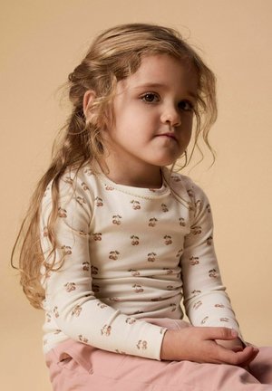 Ung jente med langt bølget hår iført en kremfarget topp med kirsebærprint og rosa bukser, sittende med hendene foldet i fanget mot en beige bakgrunn.