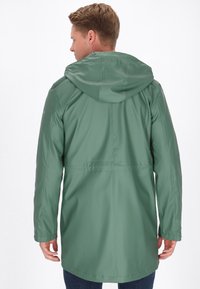 Zöld vízálló esőkabát kapucnival, hosszú ujjal és egyenes szabással. Varrott részletek és laza szabás jellemzi. A hátsó nézet látható.