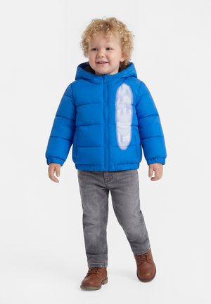 Kleinkind mit lockigem Haar, das eine blaue Steppjacke mit Kapuze, graue Jeans und braune Stiefel trägt, lächelt und geht vorwärts.