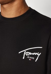 Felpa nera in cotone con colletto a costine. Presenta un logo bianco "Tommy Jeans" e un piccolo accento blu e rosso.