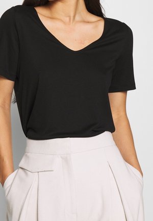 Mujer con camisa negra de manga corta y escote en V, metida por dentro de un pantalón beige claro de talle alto y pinzas, con las manos en los bolsillos.
