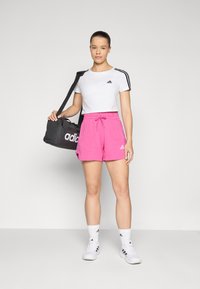 Fehér, rövidített ujjú póló fekete Adidas logóval, rózsaszín rövidnadrág és fehér cipő; egy fekete sporttáska, amelyen fehér Adidas logó található.