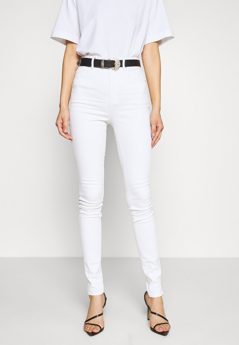 Jeans de mezclilla blanca con corte ajustado, combinados con un cinturón negro con una hebilla metálica decorativa, complementados con tacones negros con tiras.