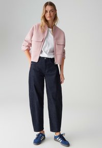 Veste courte rose avec deux poches avant, portée sur une chemise blanche. Jeans bleu marine à jambes larges et baskets bleues avec des rayures blanches.