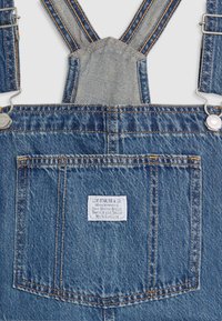Salopette en denim avec bretelles réglables, boutons en métal et une grande poche arrière avec une étiquette blanche Levi Strauss.