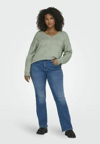 Světl zelený, oversized svetr s V výstřihem, spárovaný s modrými rovnými džíny; modelka stojí s jednou rukou v kapse a má na sobě černé boty.