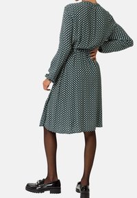 Zelené vzorované šaty s dlouhými rukávy, opaskem v pase a splývavým střihem; doplněné černými botami a průhlednými punčochami.