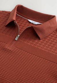 Pulover s četvrtastim zatvaranjem u rust-oranž boji napravljen od teksturiranog materijala, s rebrastim ovratnikom i horizontalnim prugama. Prednji dio s patentnim zatvaračem i metalnim produžetkom.