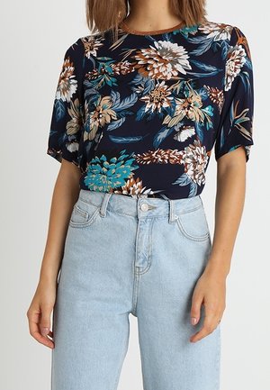 Kvinde iført en mørkeblå kortærmet bluse med stort blomsterprint, stoppet ned i lyseblå højtaljede jeans.
