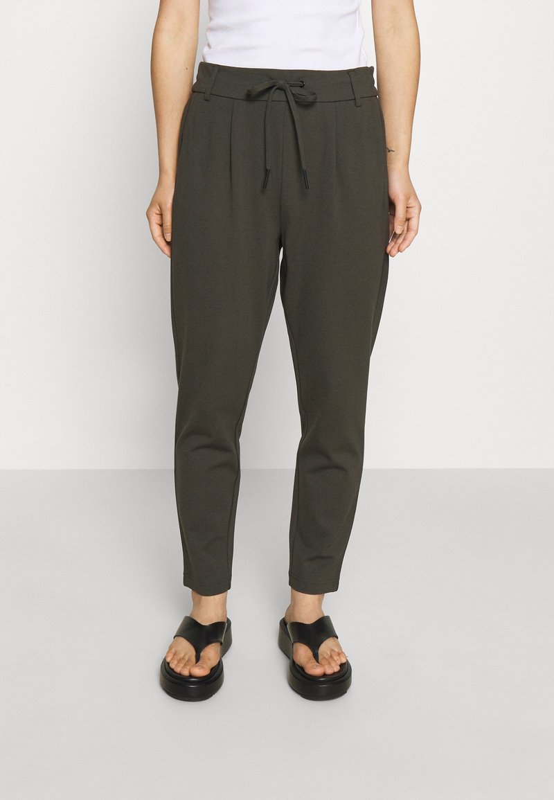 Personne portant un pantalon fuselé vert olive avec taille à cordon et des sandales plateformes noires, debout devant un fond clair uni.