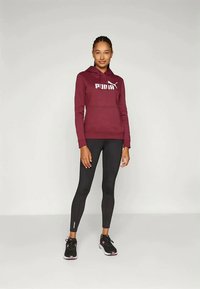 Hanorac maron cu logo alb, colanti negri si pantofi sport negri. Hanoracul are snur si buzunar cangur; colantii sunt ajustati.