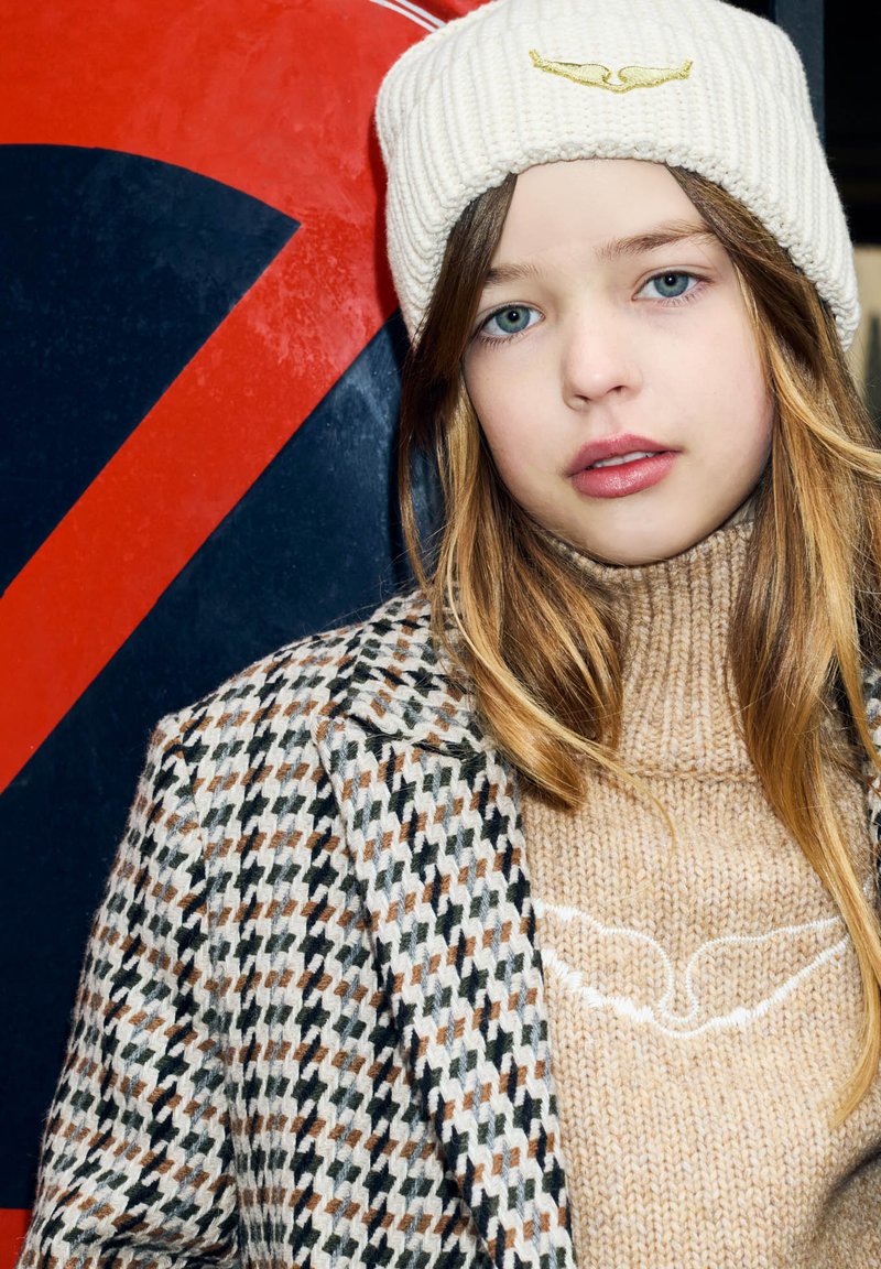 Jeune femme aux yeux bleus portant un bonnet en tricot crème et un pull col roulé sous un manteau à motifs marron, noir et blanc, se tenant devant un panneau rouge.