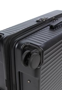 Coin de valise rigide noire avec roulettes doubles pivotantes, rainures horizontales, fermeture éclair et une poignée pliée visibles sur un fond blanc.