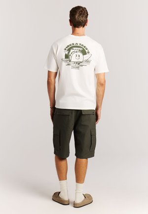 Wit katoenen T-shirt met een groene grafische print op de achterkant. Gecombineerd met olijfgroene cargoshorts, beige instapschoenen en witte sokken.