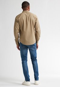 Tan lange mouw blouse met een button-down kraag, gecombineerd met blauwe jeans. De outfit heeft een relaxte pasvorm en een eenvoudig ontwerp met zichtbare stiksels.