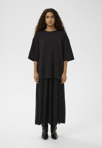 Schwarzes übergroßes T-Shirt mit kurzen Ärmeln, kombiniert mit einem langen schwarzen Rock mit transparentem Stoff, getragen mit schwarzen Stiefeletten.