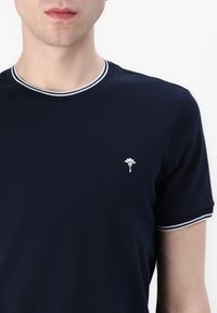 Námořnická modrá tričko s krátkým rukávem a kulatým límcem, které má bílý lem a malé bílé logo na levé části hrudi. Měkká textilie.