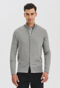 Szary sweter zapinany na zamek, wykonany z miękkiego materiału, z kołnierzem stojącym, długimi rękawami i dopasowanym krojem. Wykończony gładkim zamkiem błyskawicznym.