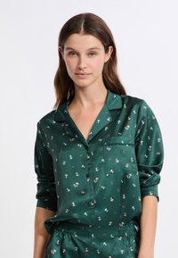 Camicia da pigiama verde in satin con tasca, caratterizzata da un motivo di funghi rosa e bianchi, con chiusura a bottoni e maniche arrotolate.