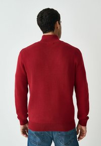 Czerwony sweter dzianinowy z wysokim kołnierzem i prążkowanymi mankietami. Materiał wydaje się gładki, dopasowany krój w jednolitym kolorze, bez widocznych wzorów.