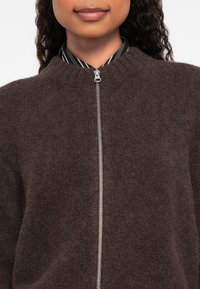 Γυναίκα φορώντας ένα σκούρο καφέ φλις μπουφάν με φερμουάρ πάνω από ένα μαύρο και άσπρο ριγωτό πουκάμισο, με τα χείλη κλειστά σε ήπιο χαμόγελο.