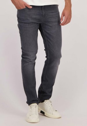 Homme portant un jean gris foncé ajusté et des baskets blanches, se tenant avec une main dans la poche devant un fond uni.