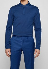 Mörkblå långärmad polotröja med krage, med en enkel knäppning i fram, tillsammans med matchande blå byxor. Slät textur överlag.