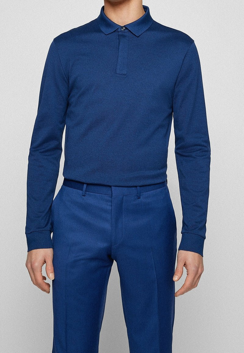 Mörkblå långärmad polotröja med krage, med en enkel knäppning i fram, tillsammans med matchande blå byxor. Slät textur överlag.
