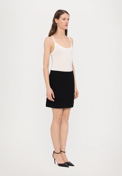 Weißer ärmelloser Bodysuit kombiniert mit einem schwarzen taillierten Minirock und schwarzen spitz zulaufenden Absätzen. Glattes Material, minimalistisches Design und Knöchelriemen.