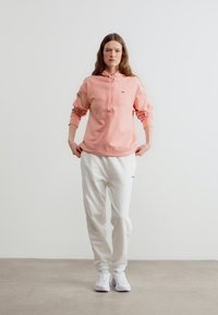 Ženska, oblečena v roza kapucarico, bele trenirke in bele superge, stoji pred enostavno svetlo podlago.