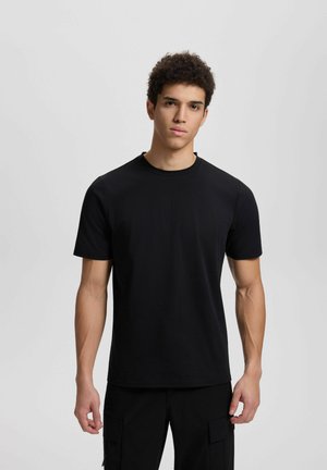 Young man with short curly hair wearing plain black short-sleeve t-shirt and black cargo pants, standing against gray background.