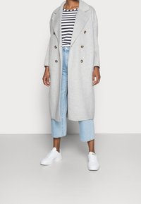 Manteau croisé gris clair avec de grands boutons, porté sur un haut à rayures noir et blanc, associé à un jean cropped bleu clair et des baskets blanches.