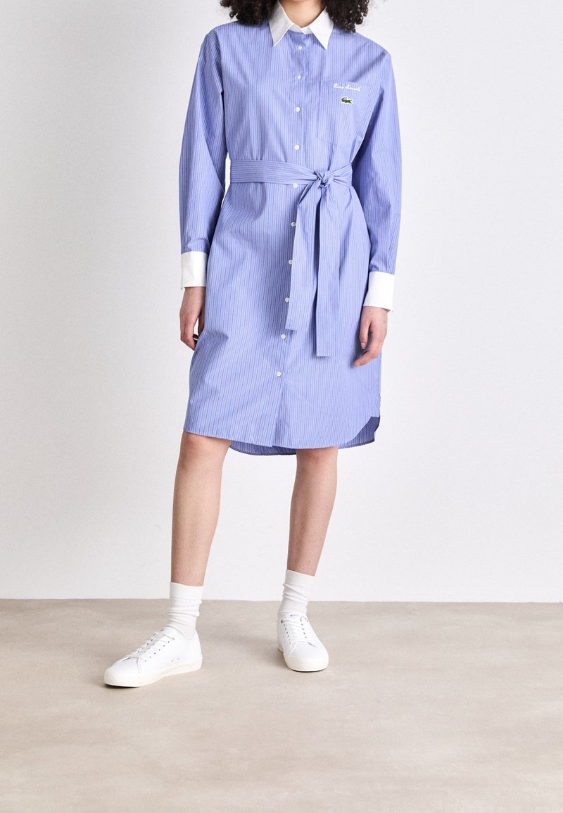 Femme portant une robe-chemise à rayures bleues avec col et poignets blancs, ceinture nouée à la taille, chaussettes blanches et baskets blanches, debout sur un sol uni.